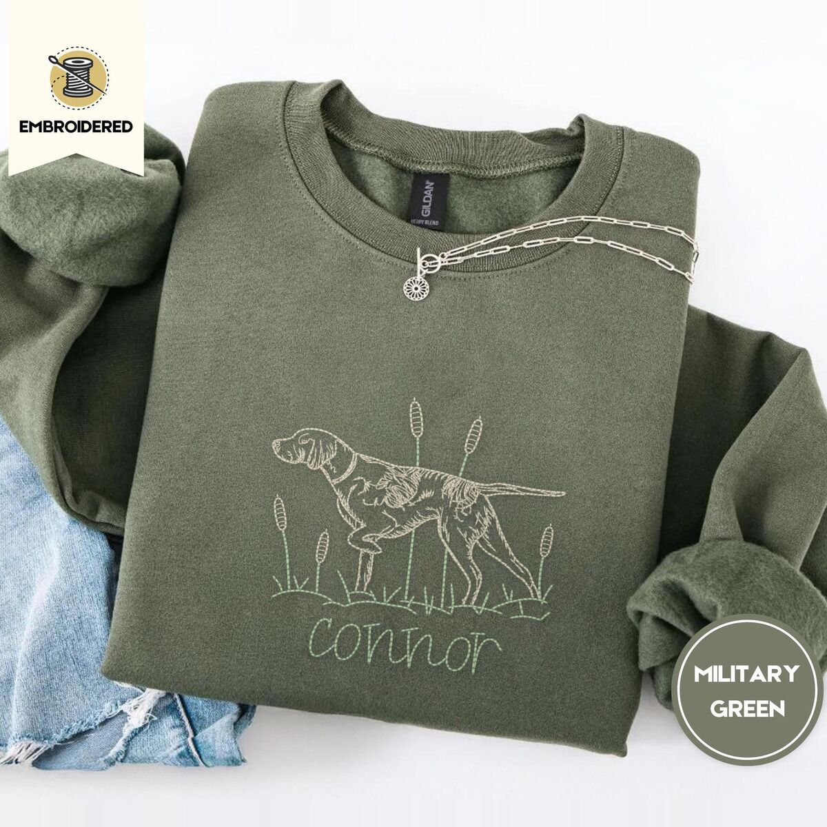 English Pointer Sweatshirt – Military Green Military Green crewneck sweatshirt folded with light jeans and a silver chain necklace, embroidered with a liver and white English pointer on point among cattails and grass, with an optional name stitched below
