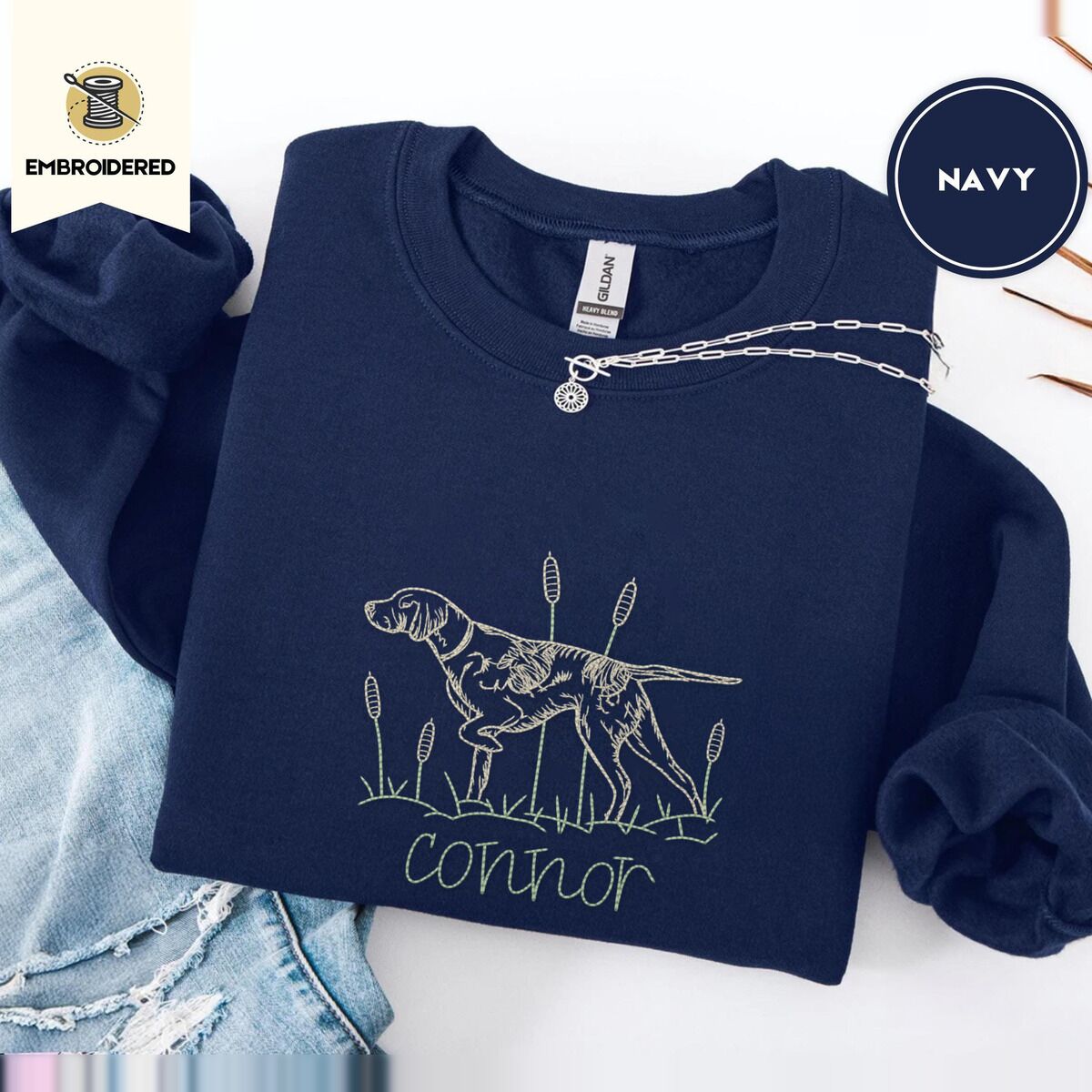 English Pointer Sweatshirt – Navy Navy crewneck sweatshirt folded with ripped jeans and a silver chain necklace, embroidered with a liver and white English pointer on point among cattails and grass, with an optional name stitched below