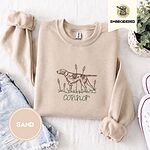Sand crewneck sweatshirt folded with denim shorts, white sneakers and a potted plant, embroidered with a liver and white English pointer on point among cattails and grass, with an optional name stitched below