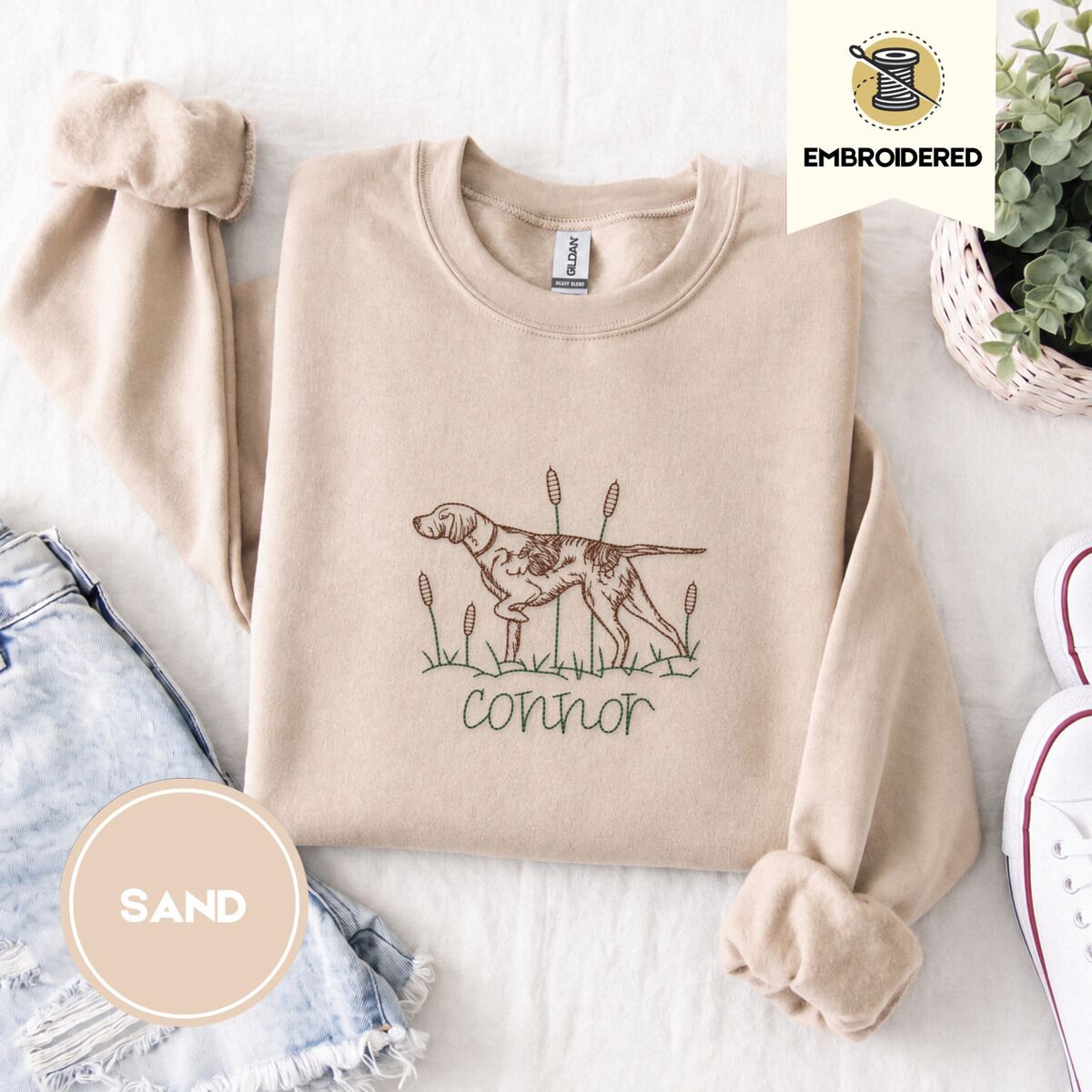 English Pointer Sweatshirt – Sand Sand crewneck sweatshirt folded with denim shorts, white sneakers and a potted plant, embroidered with a liver and white English pointer on point among cattails and grass, with an optional name stitched below