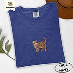 True Navy Comfort Colors T-shirt embroidered with an orange tabby Exotic Shorthair cat standing in profile with a flat face, round black eyes and a curled tail