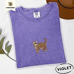 Violet Comfort Colors T-shirt embroidered with an orange tabby Exotic Shorthair cat standing in profile with a flat face, round black eyes and a curled tail