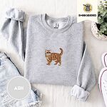 Ash Exotic Shorthair sweatshirt folded with denim shorts, white sneakers and a potted plant, with a mini embroidered orange tabby cat centered on the chest