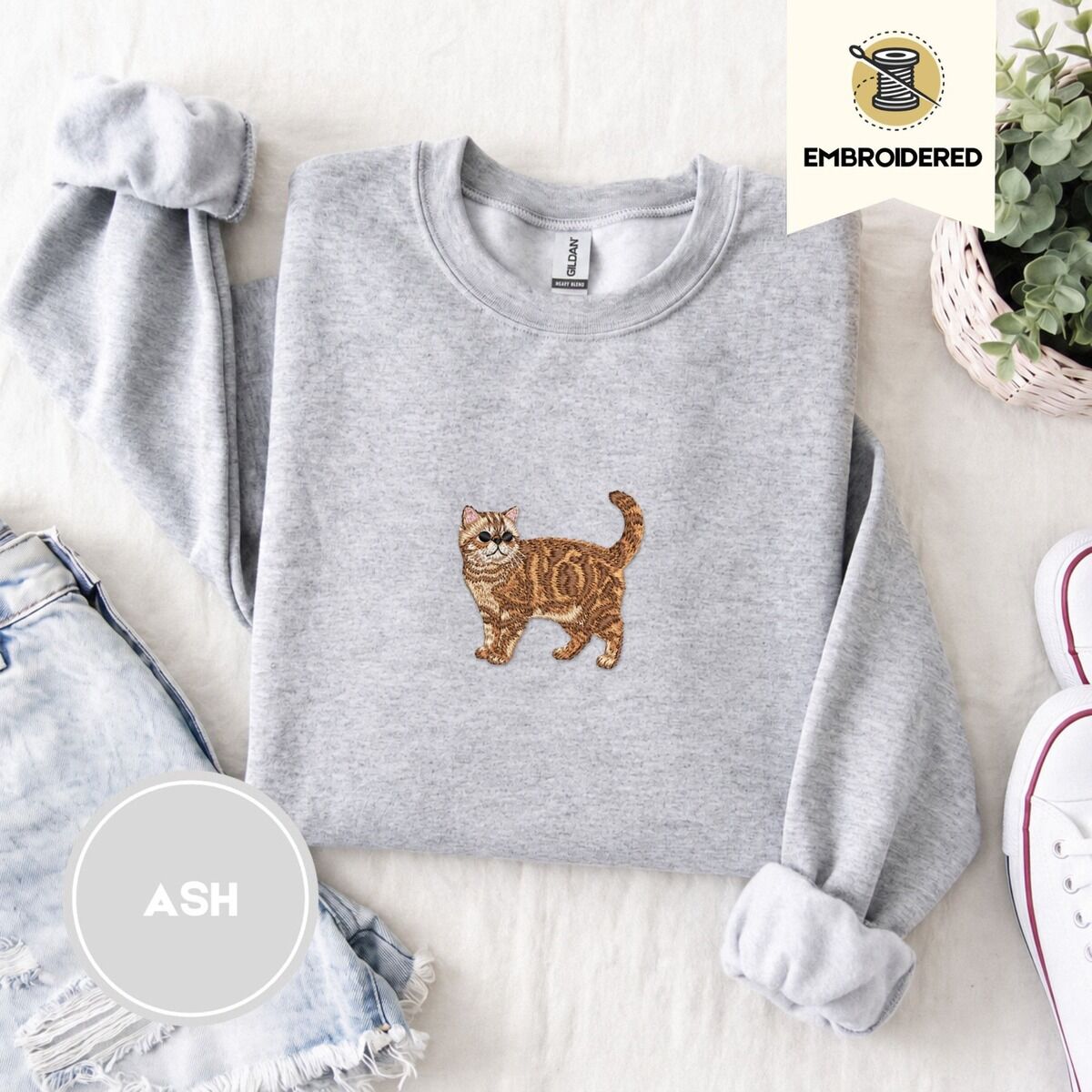 Exotic Shorthair Sweatshirt – Ash Ash Exotic Shorthair sweatshirt folded with denim shorts, white sneakers and a potted plant, with a mini embroidered orange tabby cat centered on the chest
