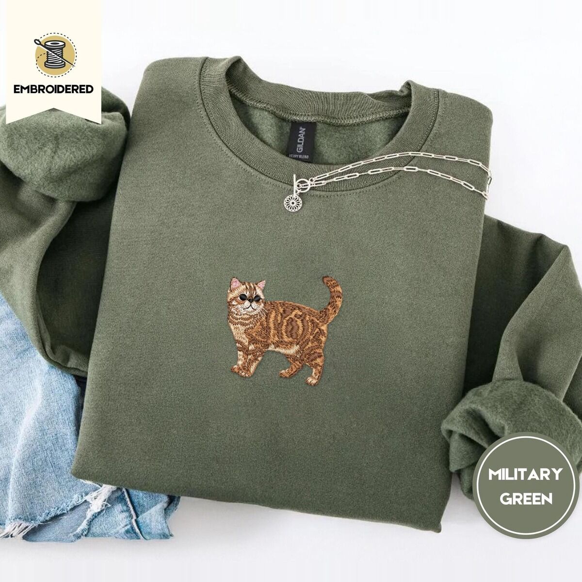 Exotic Shorthair Sweatshirt – Military Green Military Green Exotic Shorthair sweatshirt folded with light jeans and a silver chain necklace, with a mini embroidered orange tabby cat centered on the chest