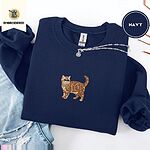 Navy Exotic Shorthair sweatshirt folded with ripped jeans and a silver chain necklace, with a mini embroidered orange tabby cat centered on the chest