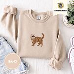 Sand Exotic Shorthair sweatshirt folded with denim shorts, white sneakers and a potted plant, with a mini embroidered orange tabby cat centered on the chest