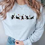 Woman in an Ash crewneck sweatshirt and jeans, with five black kittens playing in falling autumn leaves embroidered across the chest