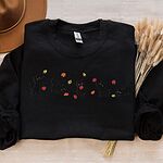 Black crewneck sweatshirt laid flat on cream linen with a brown felt hat and dried bunny tail grass, with five black kittens playing in falling autumn leaves embroidered across the chest