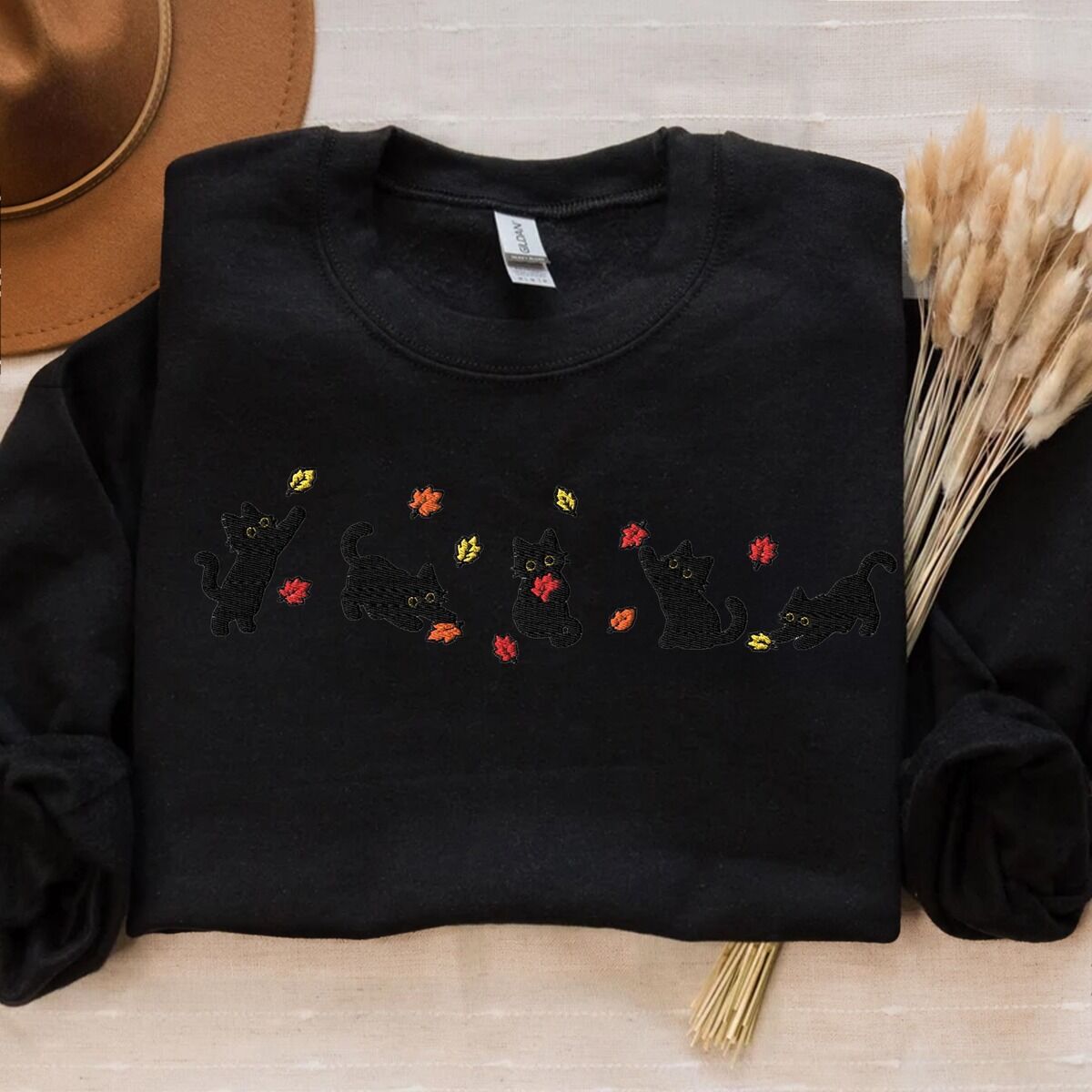 Fall Cat Sweatshirt – Black Black crewneck sweatshirt laid flat on cream linen with a brown felt hat and dried bunny tail grass, with five black kittens playing in falling autumn leaves embroidered across the chest