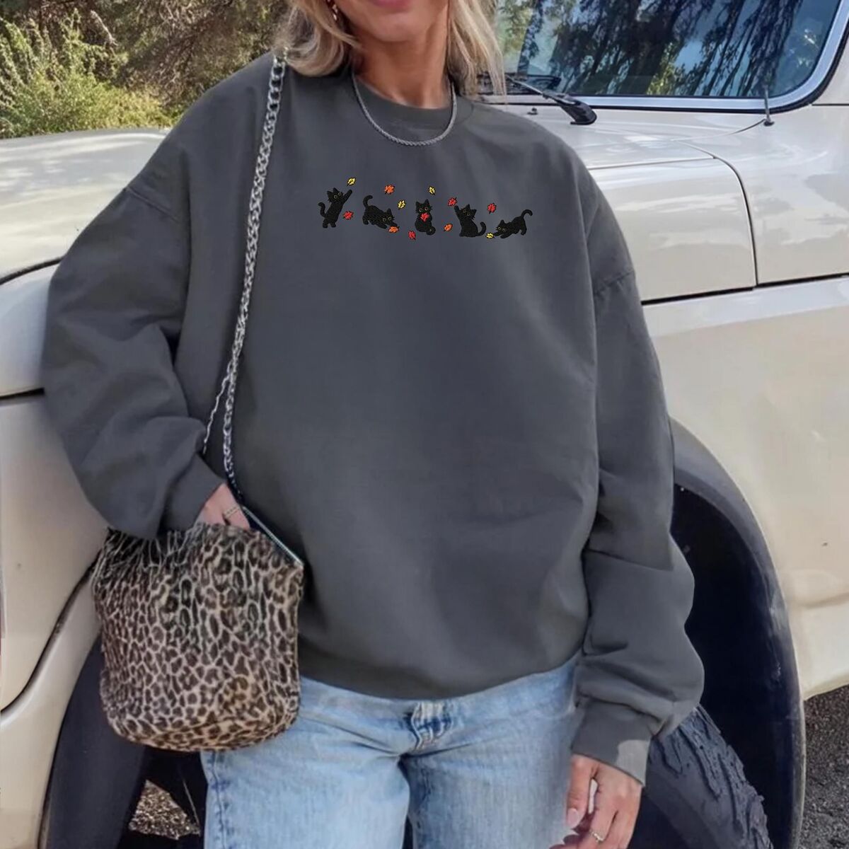 Fall Cat Sweatshirt Charcoal Woman leaning on a white pickup truck in a Charcoal crewneck sweatshirt with a leopard print bag, with five black kittens playing in falling autumn leaves embroidered across the chest