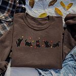 Dark Chocolate crewneck sweatshirt laid flat on a white sheet with dry leaves, a plaid shirt and jeans, with five black kittens playing in falling autumn leaves embroidered across the chest