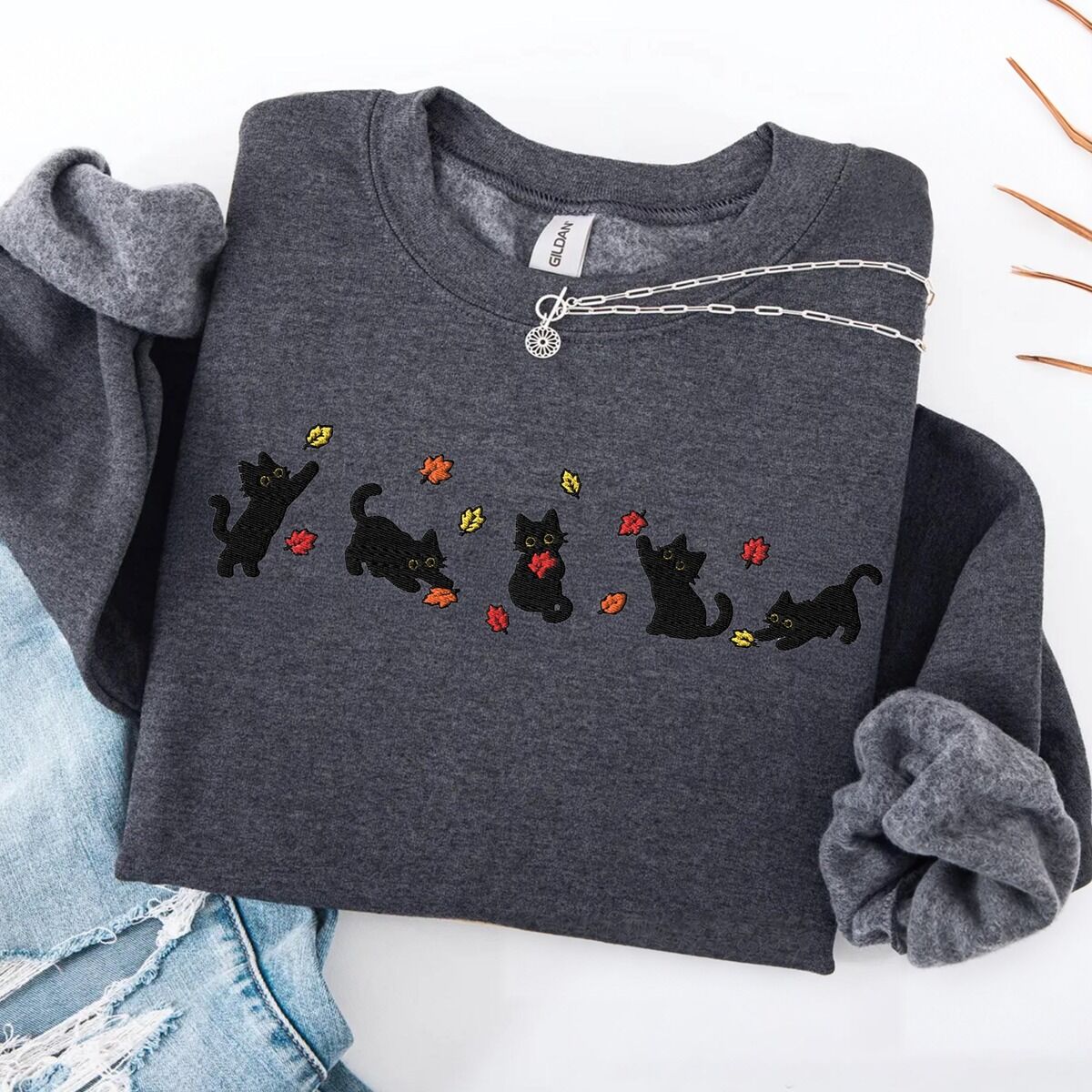 Fall Cat Sweatshirt – Dark Heather Dark Heather crewneck sweatshirt laid flat on a white table with ripped jeans and a silver necklace, with five black kittens playing in falling autumn leaves embroidered across the chest