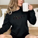 Woman sitting on a bed sipping from a white mug in a Black crewneck sweatshirt with five black kittens playing in falling autumn leaves embroidered across the chest