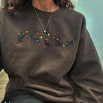 Close-up of a Dark Chocolate crewneck sweatshirt worn with a thin necklace, showing five black kittens playing in falling autumn leaves embroidered across the chest