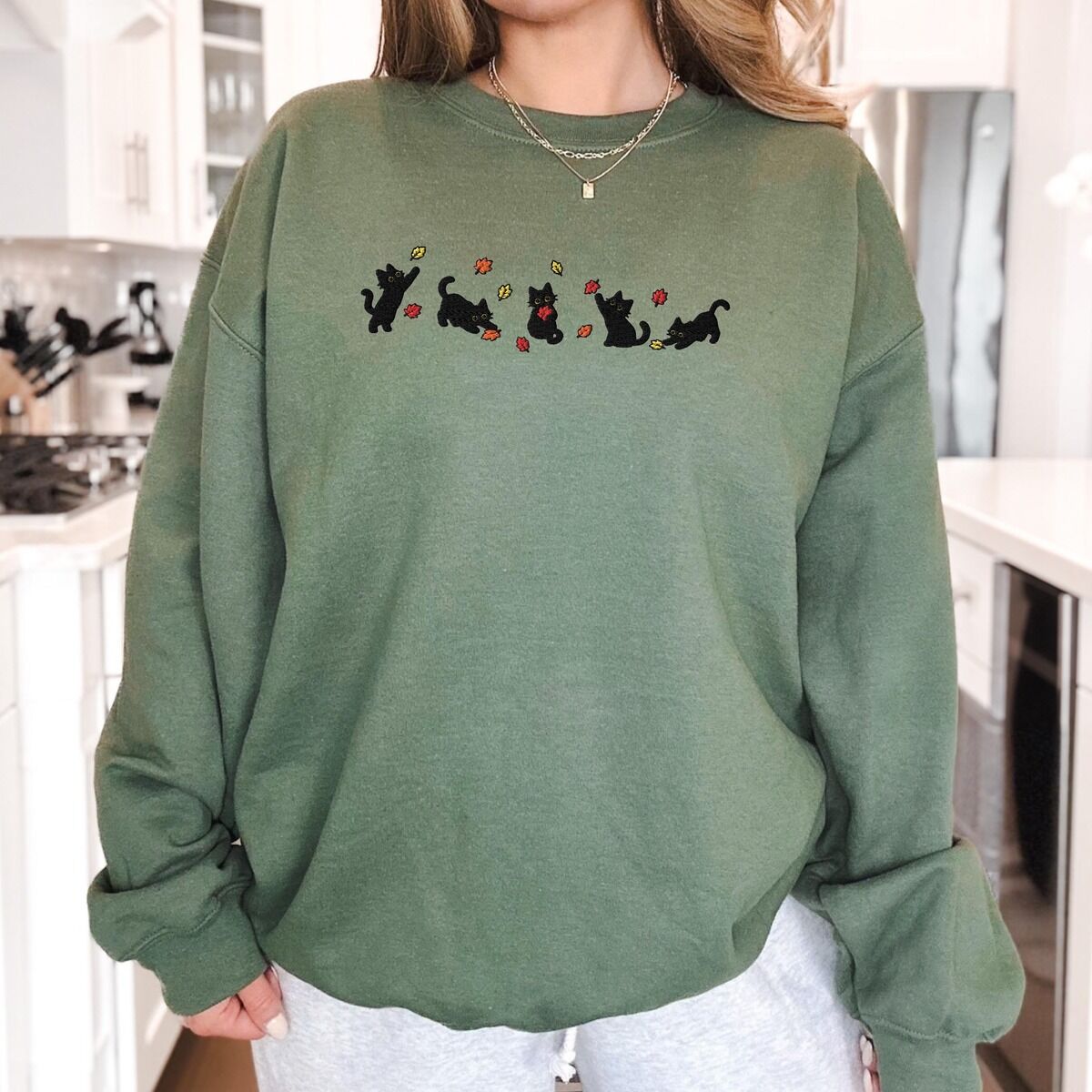 Fall Cat Sweatshirt Military Green Worn Woman standing in a kitchen in a Military Green crewneck sweatshirt with five black kittens playing in falling autumn leaves embroidered across the chest