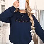 Woman in a kitchen drinking from a white mug in a Navy crewneck sweatshirt with five black kittens playing in falling autumn leaves embroidered across the chest