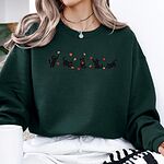 Woman sitting in a Forest Green crewneck sweatshirt holding a white mug, with five black kittens playing in falling autumn leaves embroidered across the chest