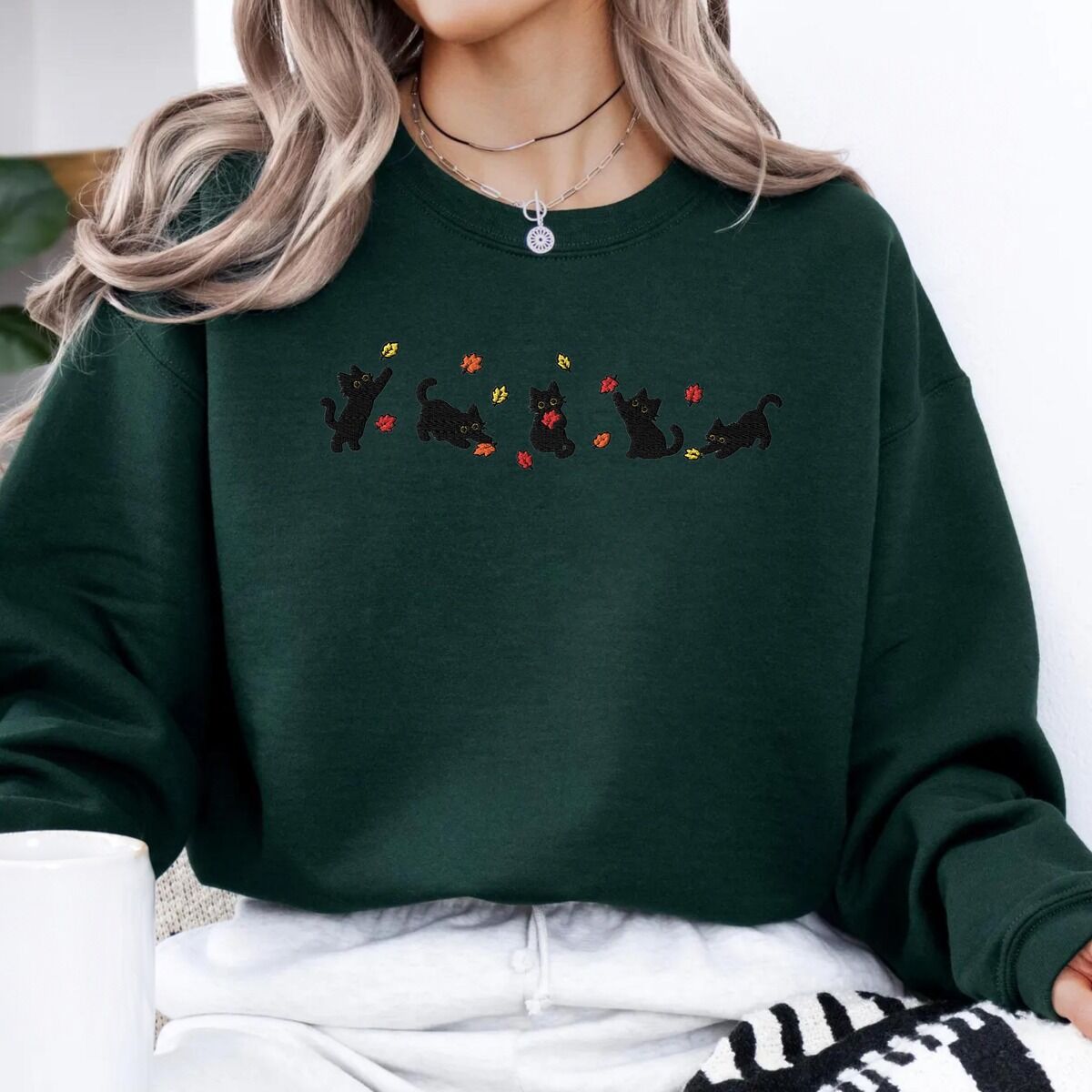Fall Cat Sweatshirt – Forest Green Woman sitting in a Forest Green crewneck sweatshirt holding a white mug, with five black kittens playing in falling autumn leaves embroidered across the chest