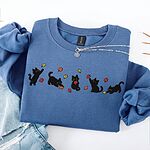 Indigo Blue crewneck sweatshirt folded on a white table with ripped jeans and a silver necklace, with five black kittens playing in falling autumn leaves embroidered across the chest