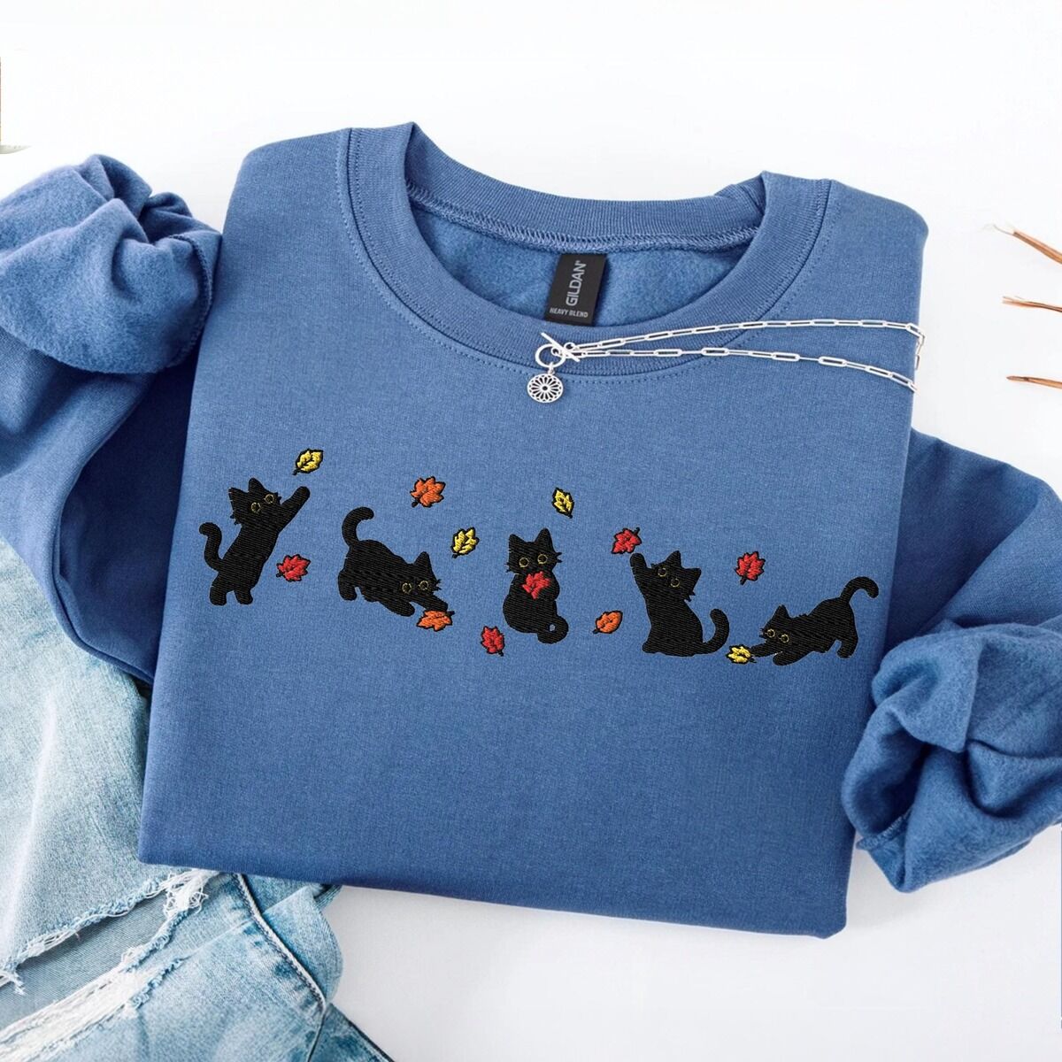 Fall Cat Sweatshirt – Indigo Blue Indigo Blue crewneck sweatshirt folded on a white table with ripped jeans and a silver necklace, with five black kittens playing in falling autumn leaves embroidered across the chest