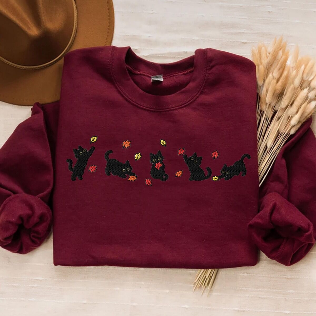 Fall Cat Sweatshirt – Maroon Maroon crewneck sweatshirt laid flat on cream linen with a brown felt hat and dried wheat, with five black kittens playing in falling autumn leaves embroidered across the chest