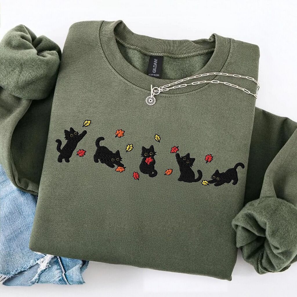 Military Green crewneck sweatshirt laid flat on a white table with ripped jeans and a silver necklace, with five black kittens playing in falling autumn leaves embroidered across the chest