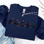 Navy crewneck sweatshirt laid flat on a white table with ripped jeans and a silver necklace, with five black kittens playing in falling autumn leaves embroidered across the chest
