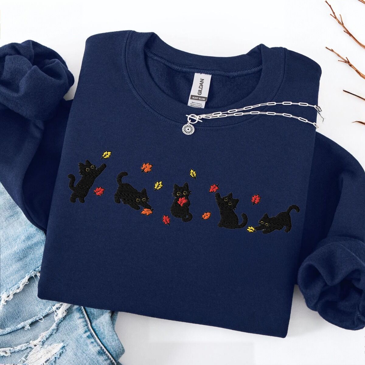 Fall Cat Sweatshirt – Navy Navy crewneck sweatshirt laid flat on a white table with ripped jeans and a silver necklace, with five black kittens playing in falling autumn leaves embroidered across the chest