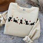 Sand crewneck sweatshirt folded on a grey stone surface next to a brown felt hat, with five black kittens playing in falling autumn leaves embroidered across the chest