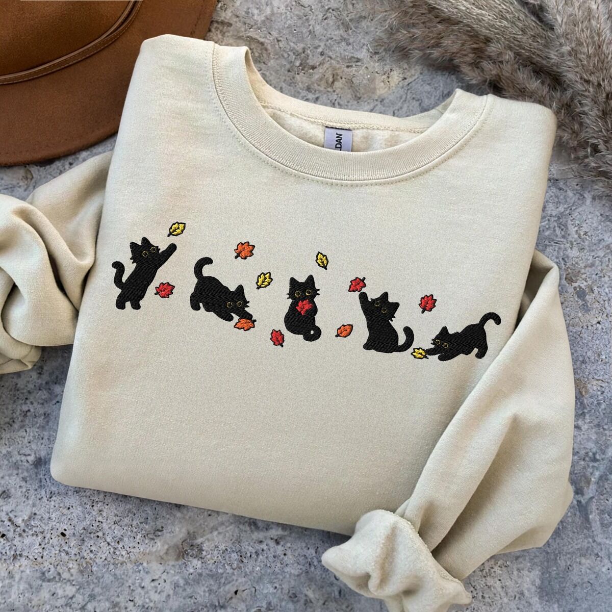 Fall Cat Sweatshirt – Sand Sand crewneck sweatshirt folded on a grey stone surface next to a brown felt hat, with five black kittens playing in falling autumn leaves embroidered across the chest