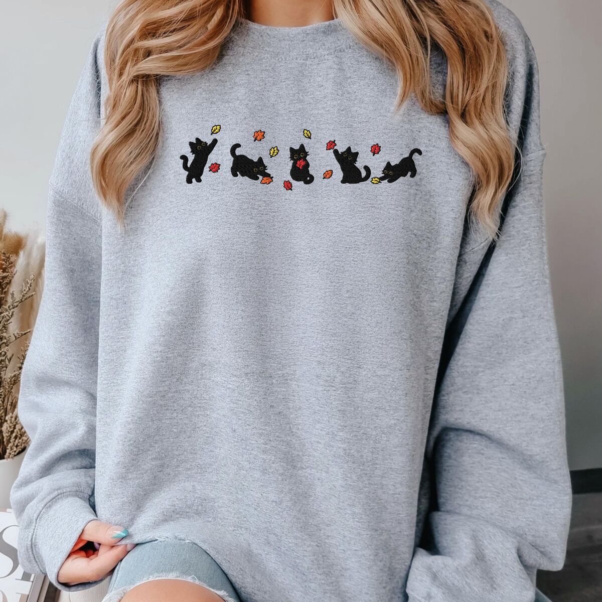 Fall Cat Sweatshirt Sport Grey Woman sitting in a Sport Grey crewneck sweatshirt next to dried pampas grass, with five black kittens playing in falling autumn leaves embroidered across the chest