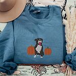 Indigo Blue crewneck sweatshirt laid flat on a patterned throw with a felt hat and pampas grass, embroidered with a black and white tuxedo pit bull sitting between two orange pumpkins