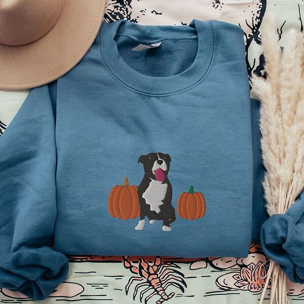 Indigo Blue crewneck sweatshirt laid flat on a patterned throw with a felt hat and pampas grass, embroidered with a black and white tuxedo pit bull sitting between two orange pumpkins