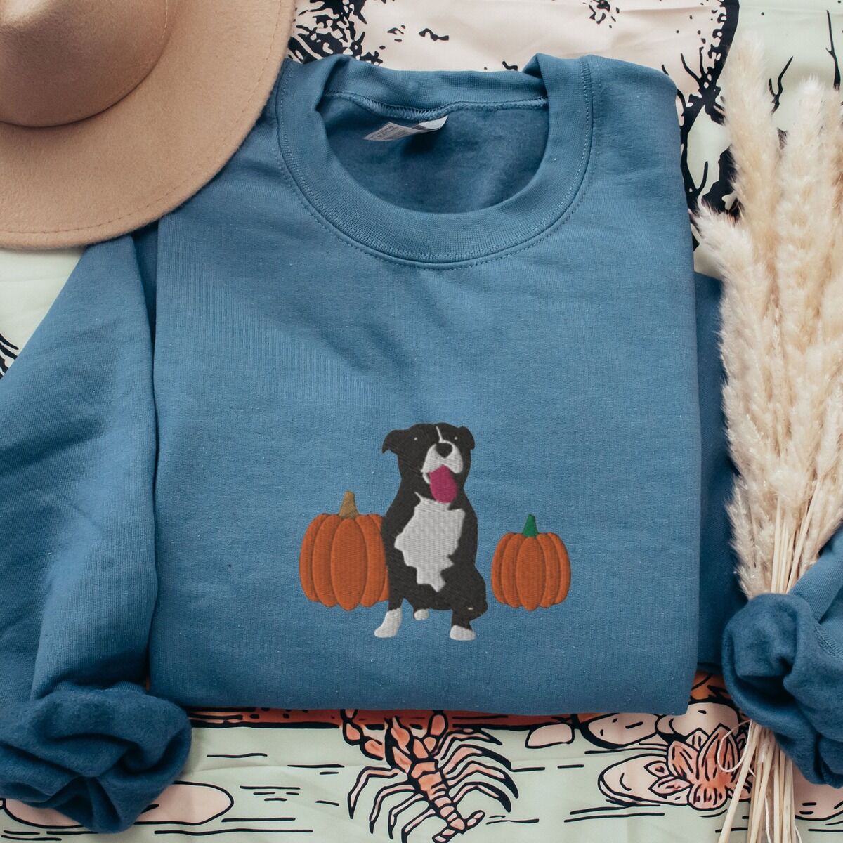 Fall Pitbull Sweatshirt – Indigo Blue Indigo Blue crewneck sweatshirt laid flat on a patterned throw with a felt hat and pampas grass, embroidered with a black and white tuxedo pit bull sitting between two orange pumpkins