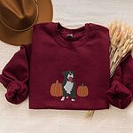 Maroon crewneck sweatshirt laid flat with a felt hat and dried wheat, embroidered with a black and white tuxedo pit bull sitting between two orange pumpkins
