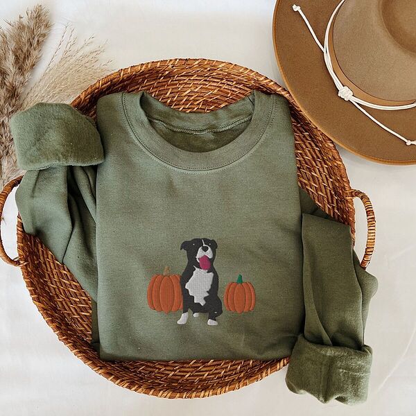 Military Green crewneck sweatshirt folded in a woven basket next to a felt hat, embroidered with a black and white tuxedo pit bull sitting between two orange pumpkins
