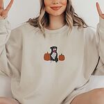 Smiling woman making peace signs in a Sand crewneck sweatshirt, embroidered with a black and white tuxedo pit bull sitting between two orange pumpkins