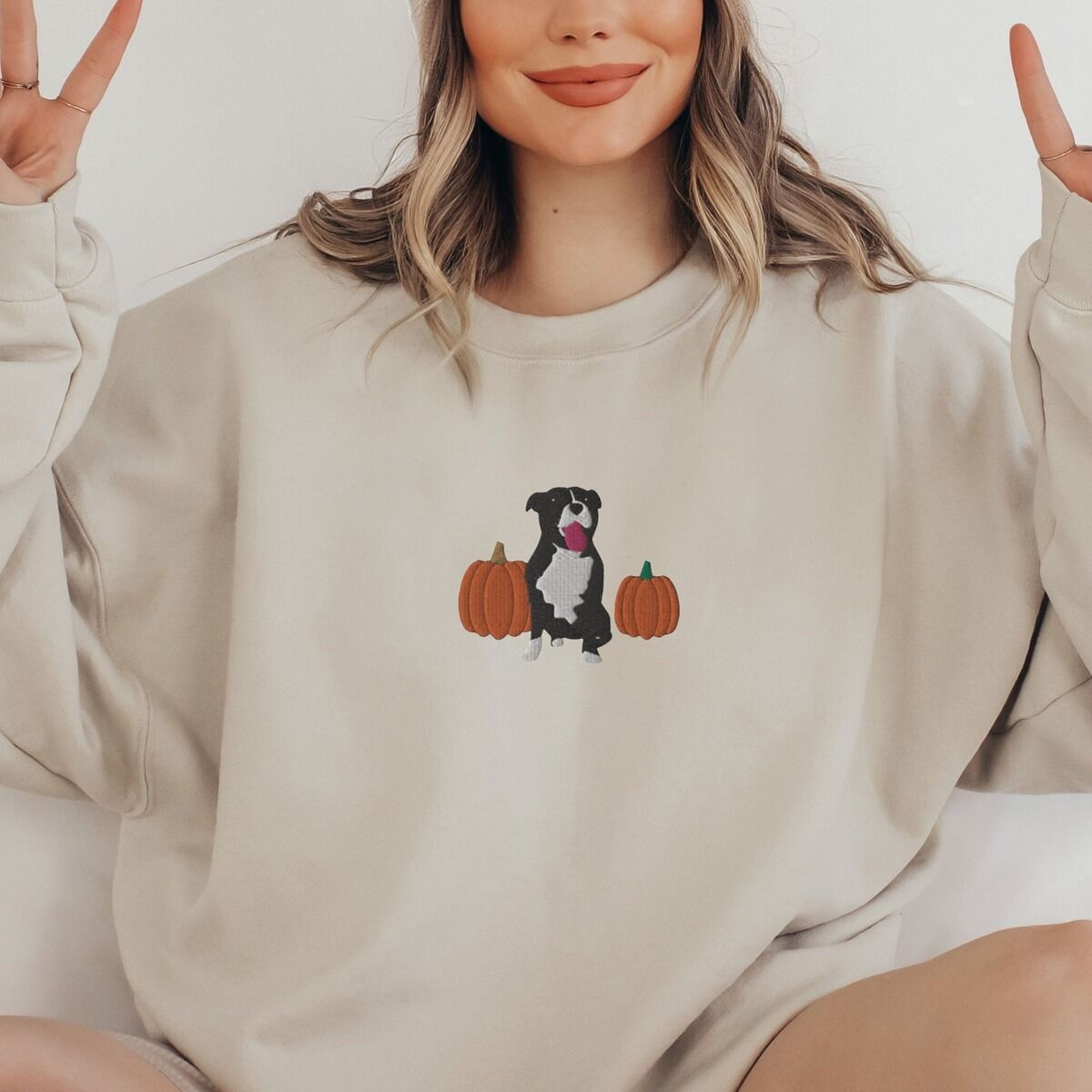 Fall Pitbull Sweatshirt – Sand Smiling woman making peace signs in a Sand crewneck sweatshirt, embroidered with a black and white tuxedo pit bull sitting between two orange pumpkins