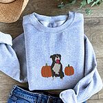 Sport Grey crewneck sweatshirt laid flat on a wood floor with jeans, a straw hat and greenery, embroidered with a black and white tuxedo pit bull sitting between two orange pumpkins