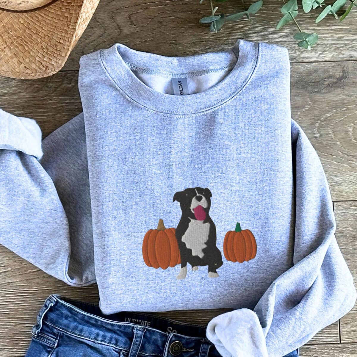 Fall Pitbull Sweatshirt - Sport Grey Sport Grey crewneck sweatshirt laid flat on a wood floor with jeans, a straw hat and greenery, embroidered with a black and white tuxedo pit bull sitting between two orange pumpkins