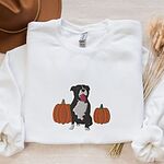 White crewneck sweatshirt laid flat with a felt hat and dried wheat, embroidered with a black and white tuxedo pit bull sitting between two orange pumpkins