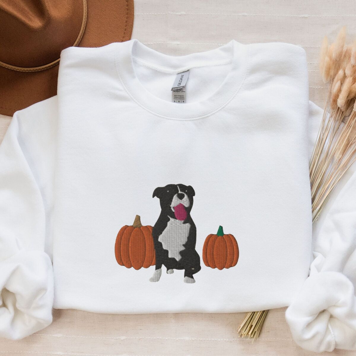 Fall Pitbull Sweatshirt - White White crewneck sweatshirt laid flat with a felt hat and dried wheat, embroidered with a black and white tuxedo pit bull sitting between two orange pumpkins