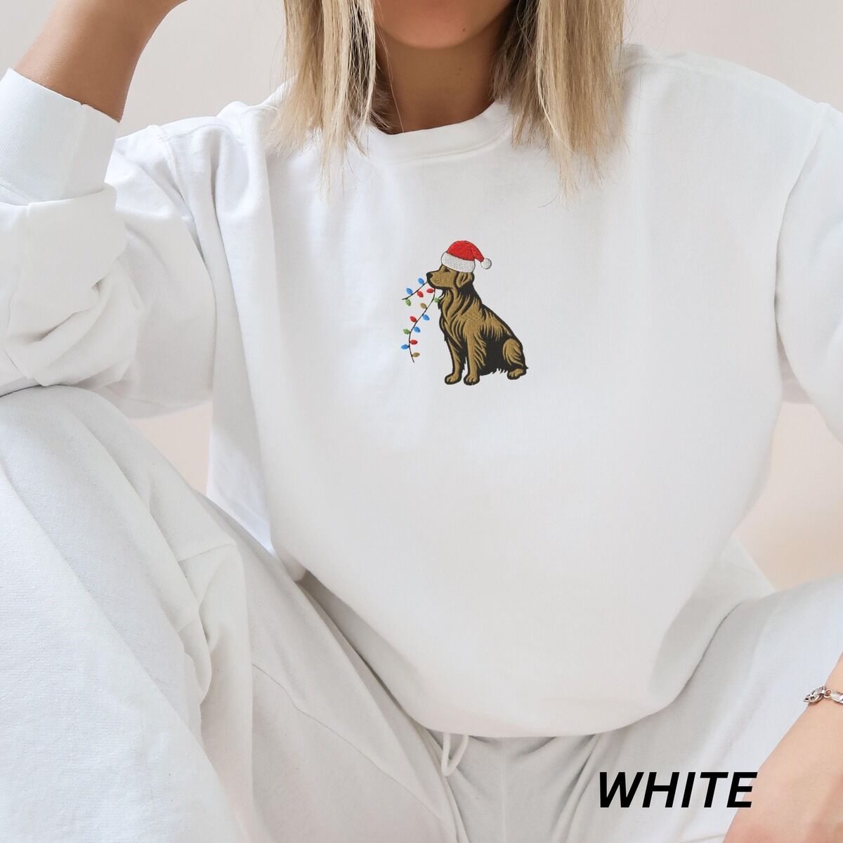 Festive Golden Retriever Sweatshirt – White Woman sitting in a White crewneck sweatshirt embroidered with a golden retriever in a red Santa hat carrying colored lights