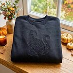 Black crewneck sweatshirt folded on a wooden windowsill beside mini pumpkins, a candle and wildflowers, embroidered black on black with a sitting cat silhouette filled with flowers and leaves