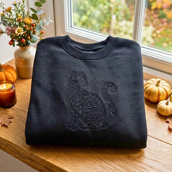 Black crewneck sweatshirt folded on a wooden windowsill beside mini pumpkins, a candle and wildflowers, embroidered black on black with a sitting cat silhouette filled with flowers and leaves