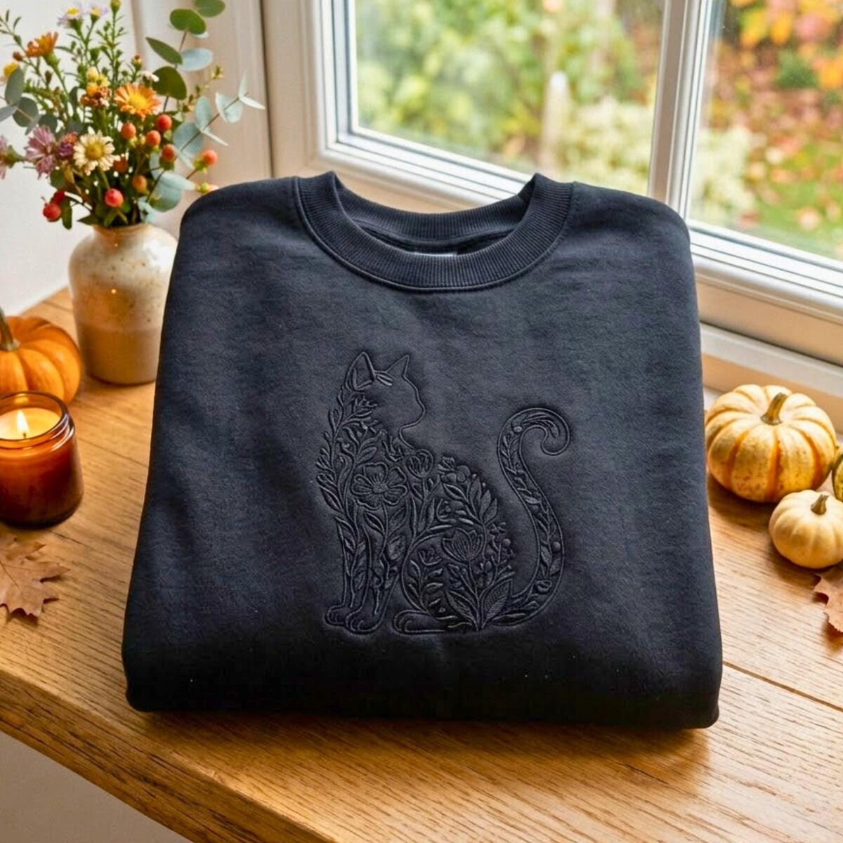 Floral Black Cat Sweatshirt – Black Black crewneck sweatshirt folded on a wooden windowsill beside mini pumpkins, a candle and wildflowers, embroidered black on black with a sitting cat silhouette filled with flowers and leaves