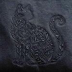 Close-up of the black-on-black embroidery: a sitting cat outline filled with wildflowers, leaves and berries, with its tail curled into a swirl
