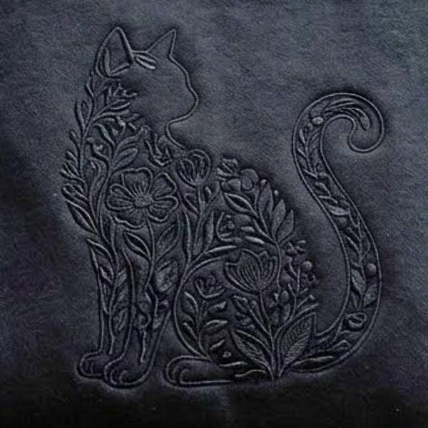 Close-up of the black-on-black embroidery: a sitting cat outline filled with wildflowers, leaves and berries, with its tail curled into a swirl