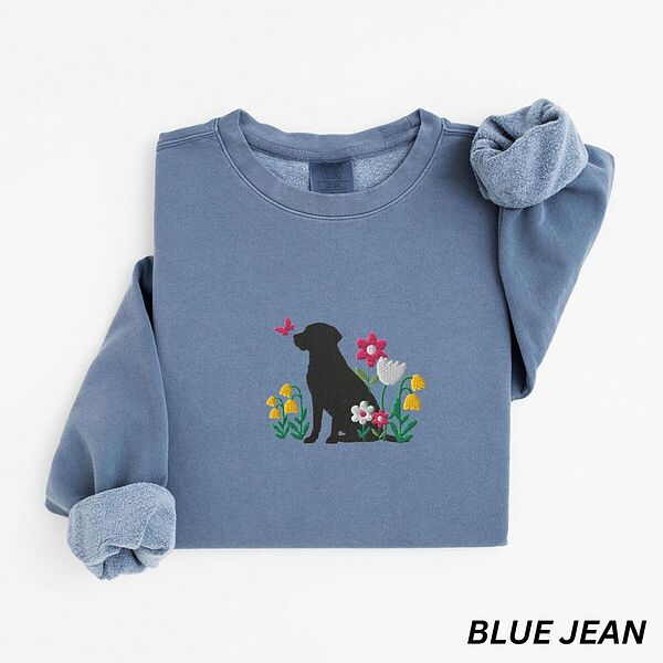 Blue Jean crewneck sweatshirt laid flat on white, embroidered with a sitting black Labrador silhouette among yellow bell flowers, a white tulip and pink daisies, with a pink butterfly